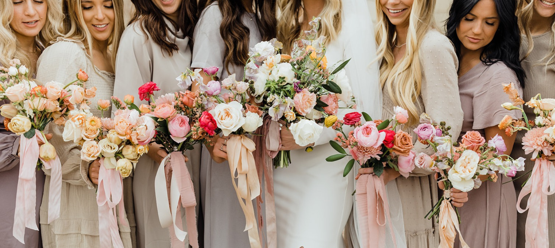 A group of bridesmaids and a bride holding matching bouquets of pink, peach, white, and green flowers with long satin ribbons.