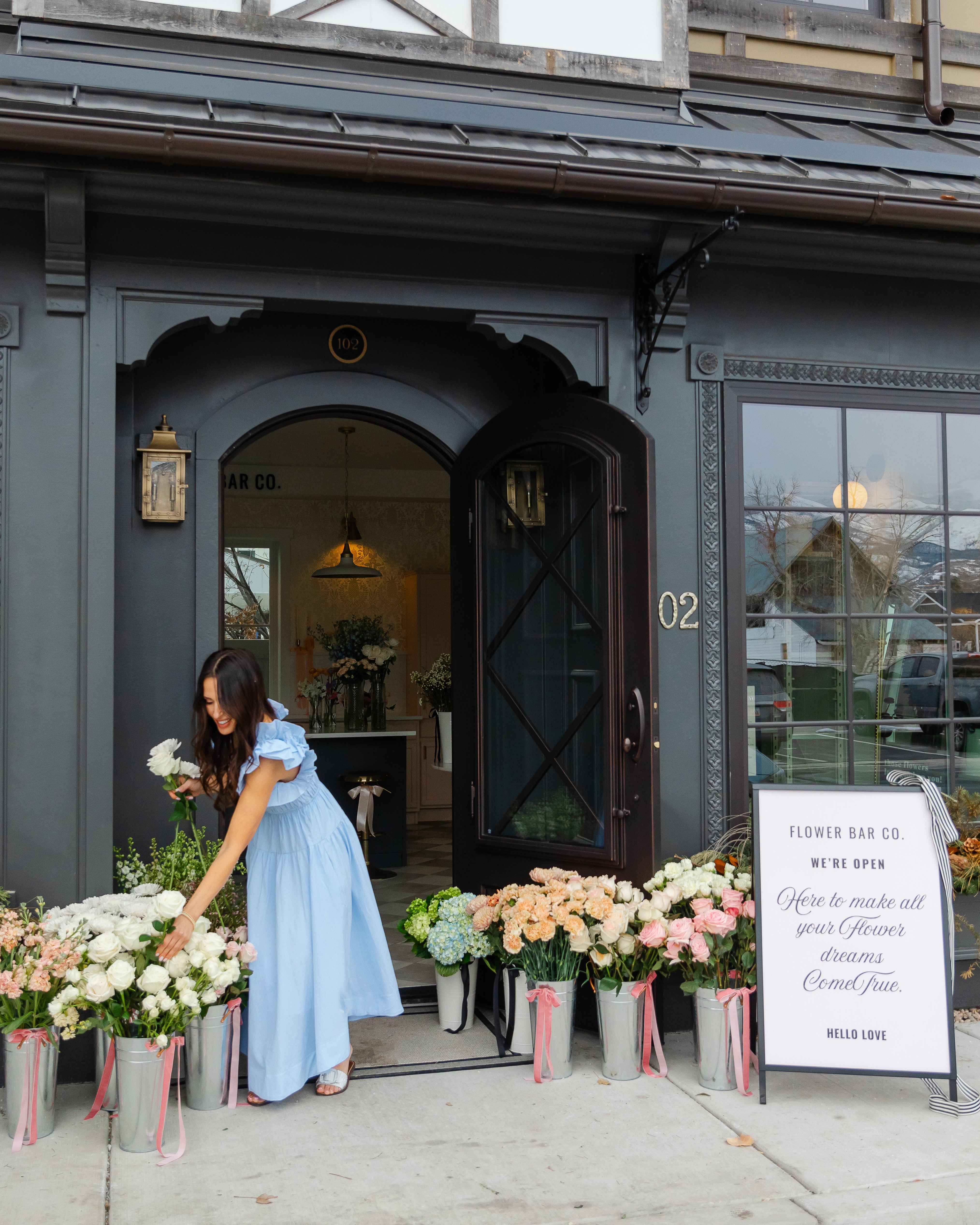 Woman in a blue dress arranging flowers outside a building with a sign.