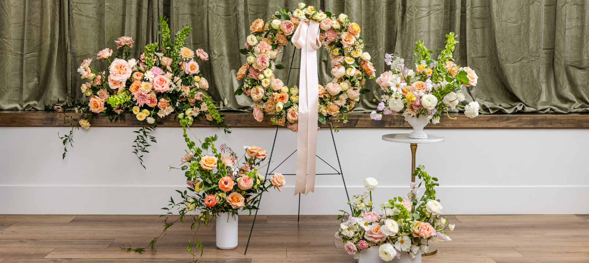 A sympathy floral arrangement display featuring a large arch and multiple bouquets in soft peach, pink, cream, and white tones.