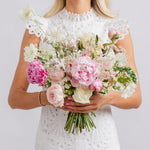 A bride holding a large bridal bouquet of pink and white flowers, including peonies, roses, and white chrysanthemums.