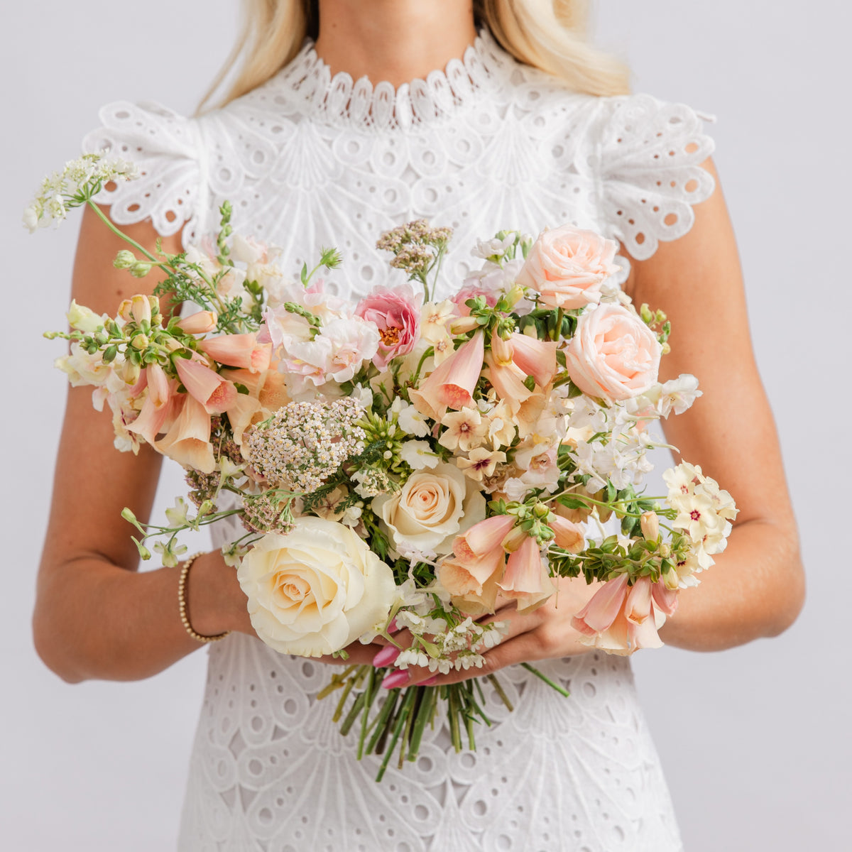 A bride holding a large bridal bouquet of pale pink, cream, and white flowers with green stems.