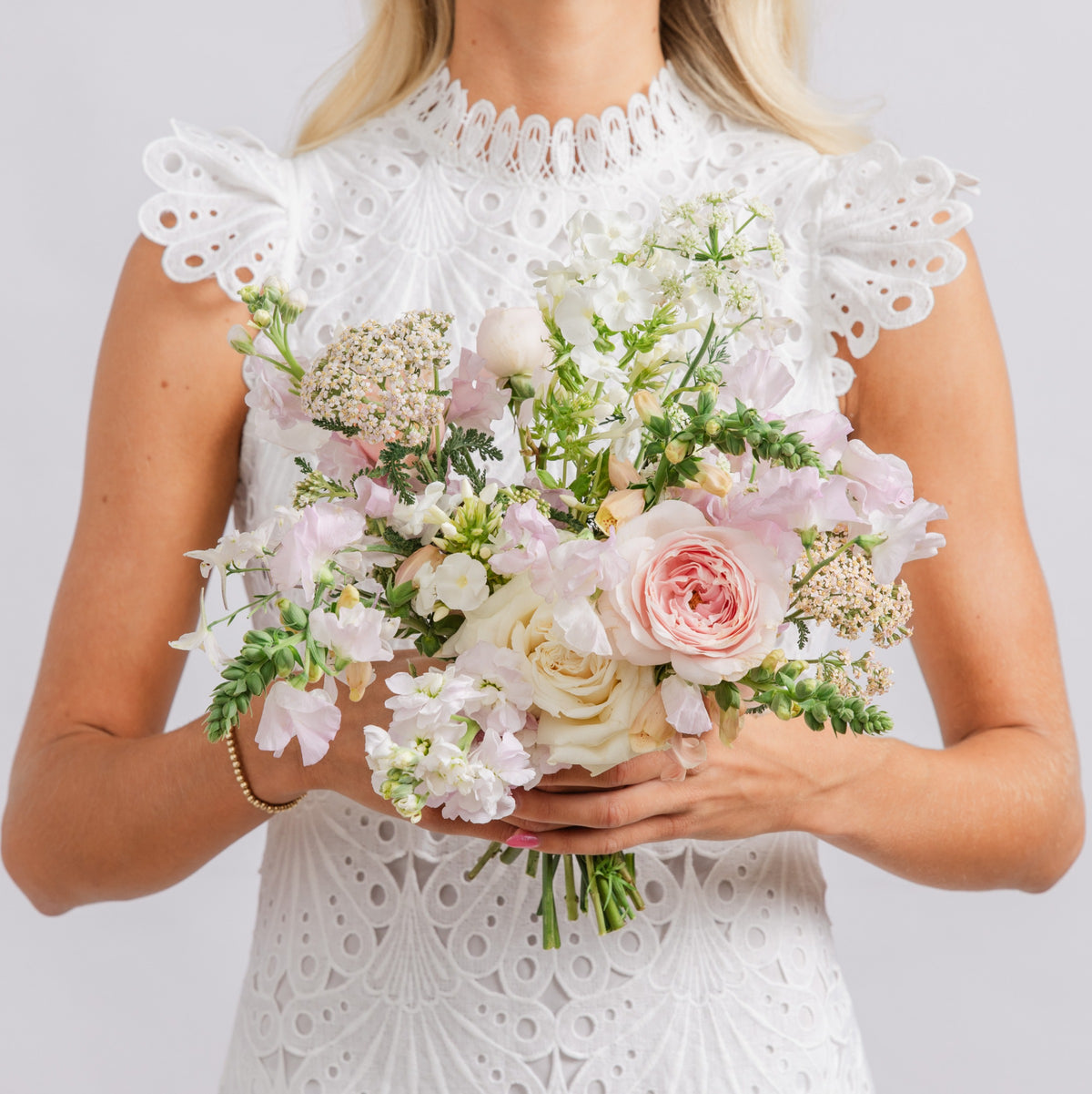 A bride holding a soft pastel wedding bouquet with pink and cream flowers and green foliage.