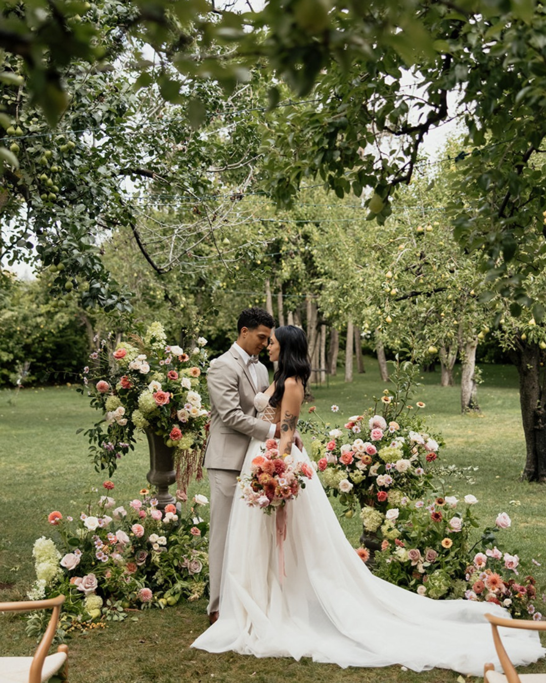 A bride in a long white wedding gown holding a pink bouquet stands beside a groom in a light gray suit, embracing outdoors on a grassy lawn decorated with large floral arrangements and trees.