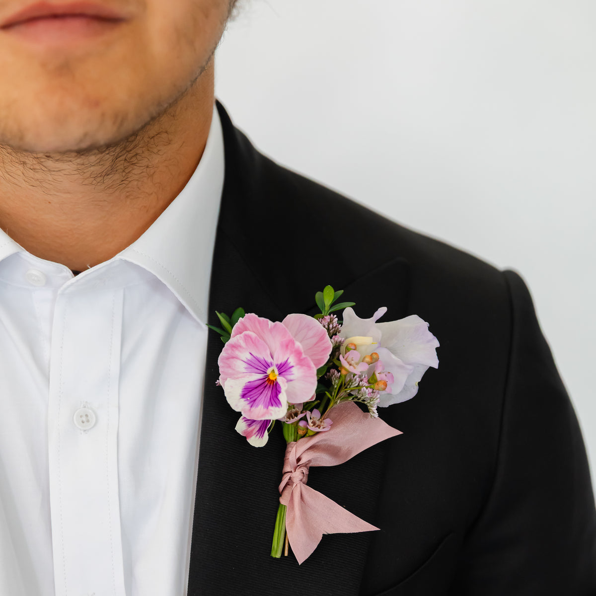 A black suit jacket and white dress shirt, featuring a pink and purple flower dance or wedding boutonniere on the left chest with a dusty pink ribbon.
