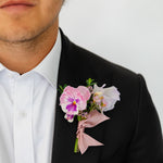A black suit jacket and white dress shirt, featuring a pink and purple flower dance or wedding boutonniere on the left chest with a dusty pink ribbon.