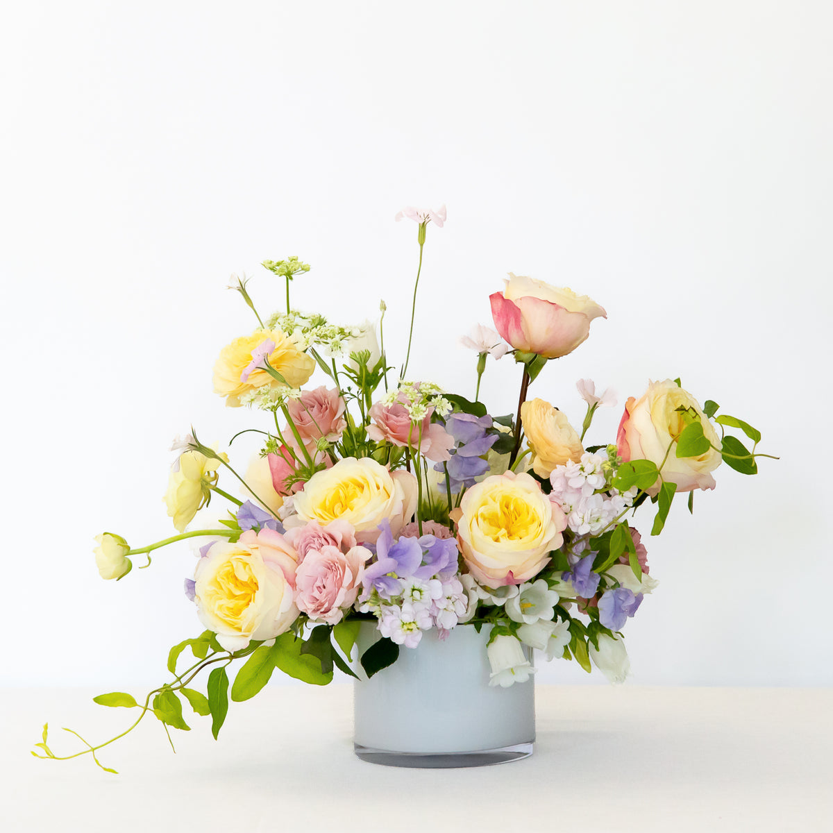 A large, lush, pastel flower bouquet in a white glass vase, featuring yellow, pink, and peach roses with green foliage.