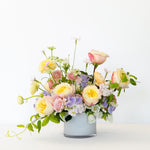 A large, lush, pastel flower bouquet in a white glass vase, featuring yellow, pink, and peach roses with green foliage.