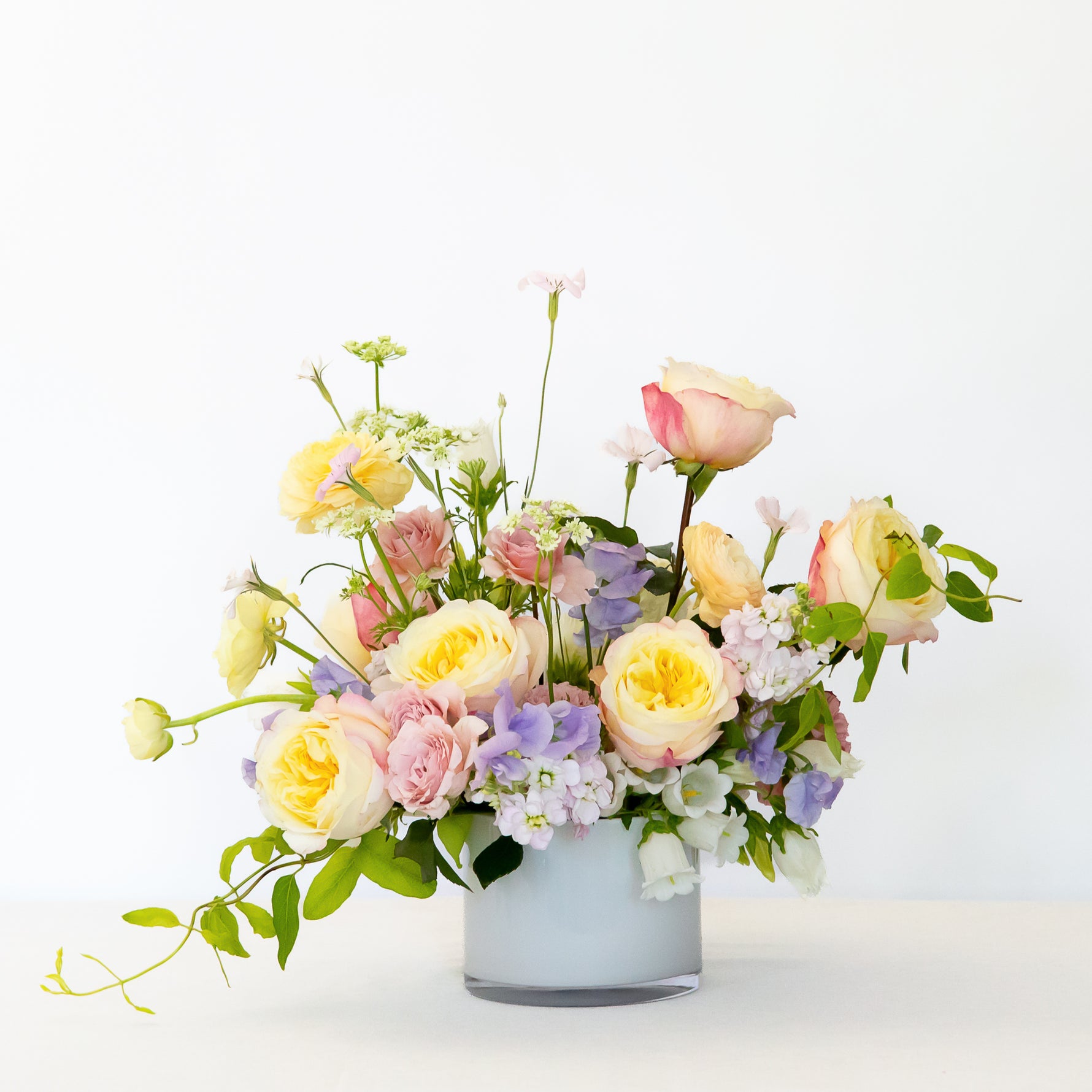 A large, lush, pastel flower bouquet in a white glass vase, featuring yellow, pink, and peach roses with green foliage.
