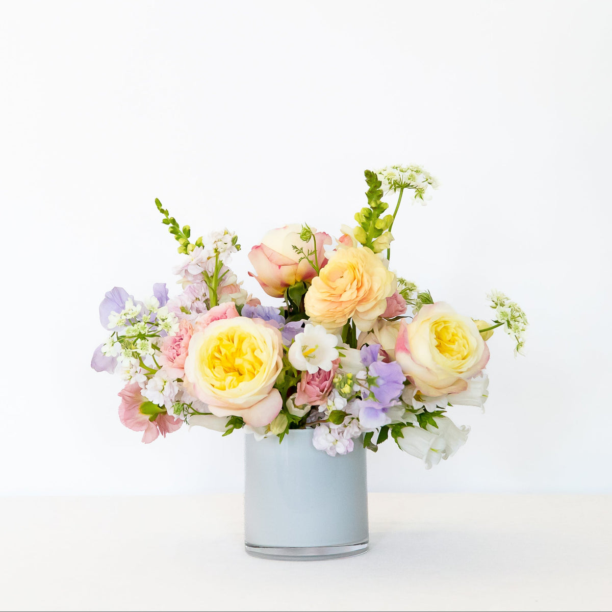 A pastel flower arrangement in a white glass vase, featuring pale yellow roses, peach roses, lavender sprigs, and white filler flowers.