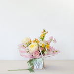 A small floral bouquet wrapped in pink striped paper and tied with a green ribbon, presented in a square box with a card that reads 'FLOWER'. The bouquet contains pale yellow, pink, and white flowers with green stems.