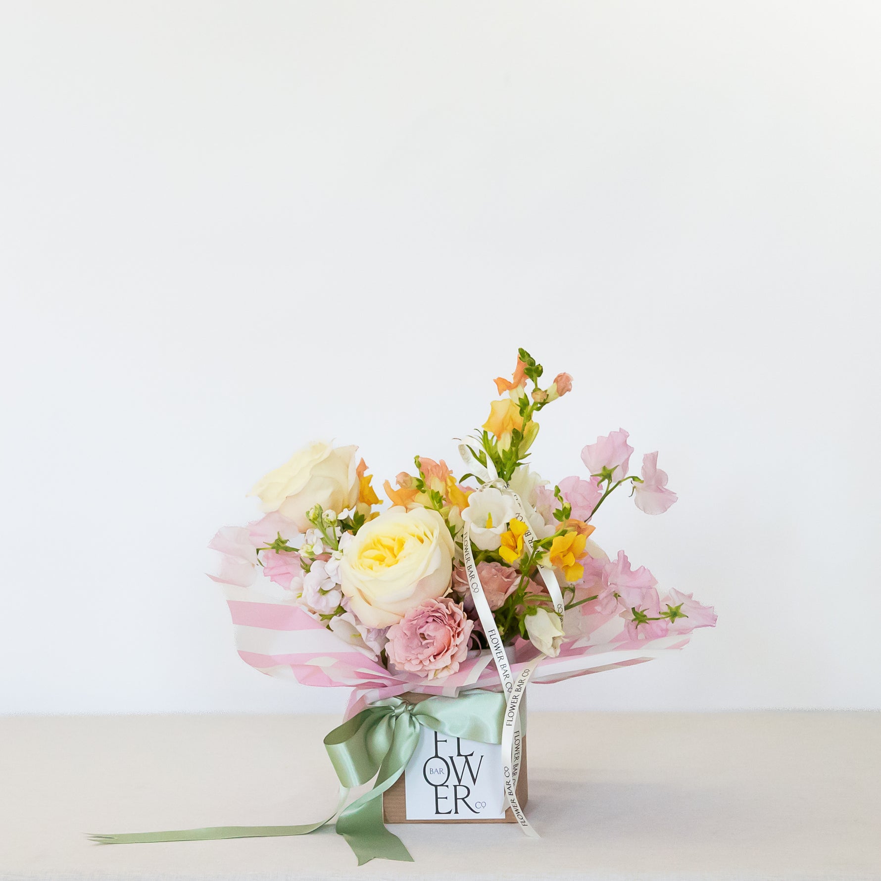 A small floral bouquet wrapped in pink striped paper and tied with a green ribbon, presented in a square box with a card that reads 'FLOWER'. The bouquet contains pale yellow, pink, and white flowers with green stems.