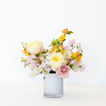 A pastel flower bouquet in a frosted glass vase, featuring cream roses, pink carnations, yellow snapdragons, and white blossoms.