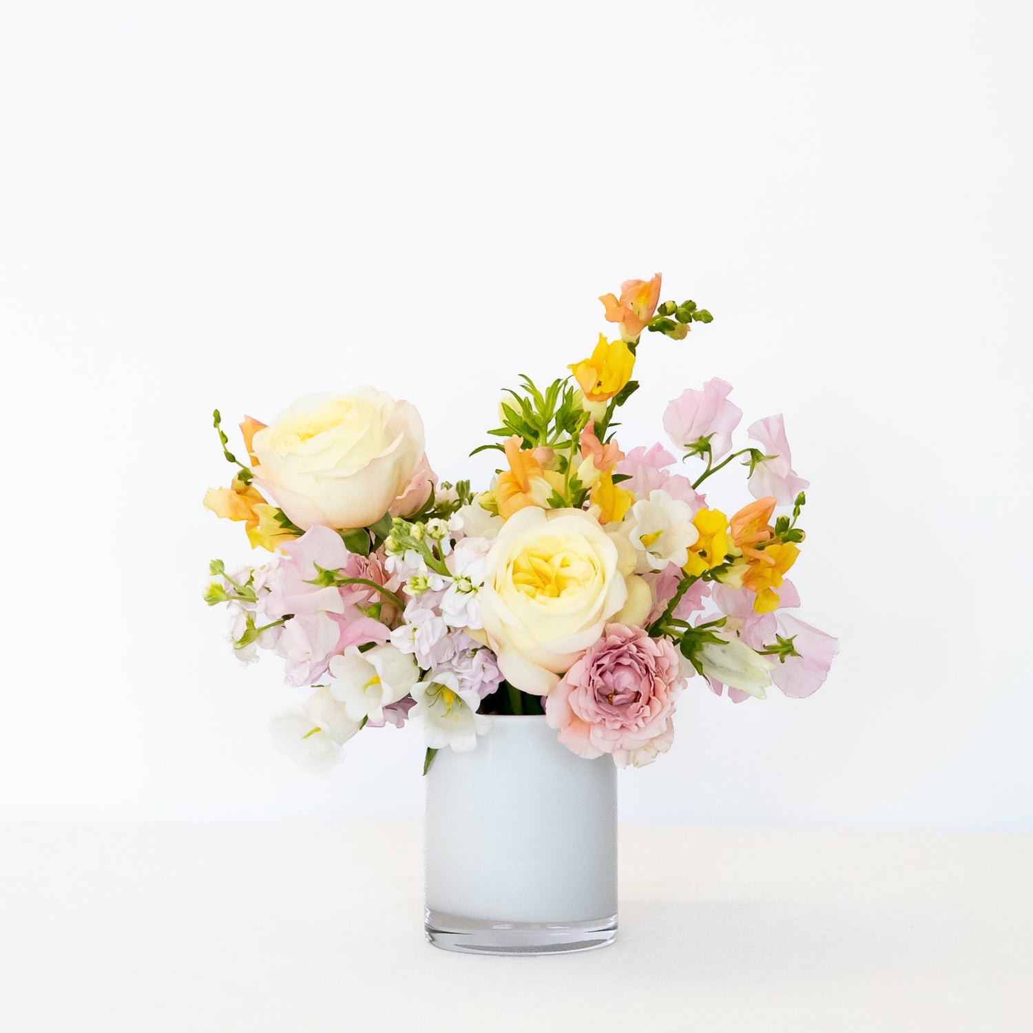 A pastel flower bouquet in a frosted glass vase, featuring cream roses, pink carnations, yellow snapdragons, and white blossoms.