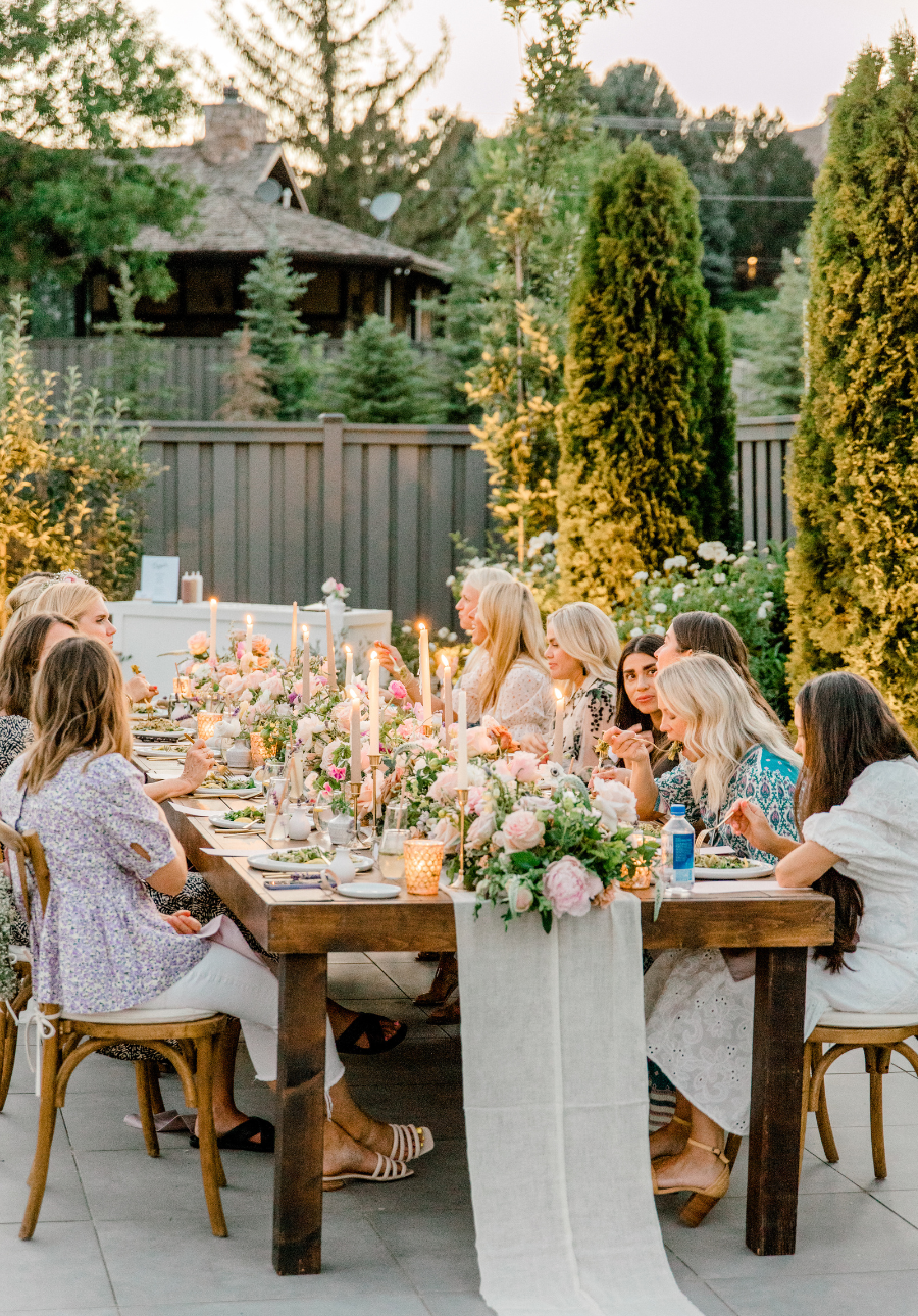 A long wooden dining table set for an outdoor patio dinner party in Utah, surrounded by guests seated in wooden chairs. The table is overflowing with lush pink and white floral arrangements, lit candles, and a white runner, with a garden and fence in the background.