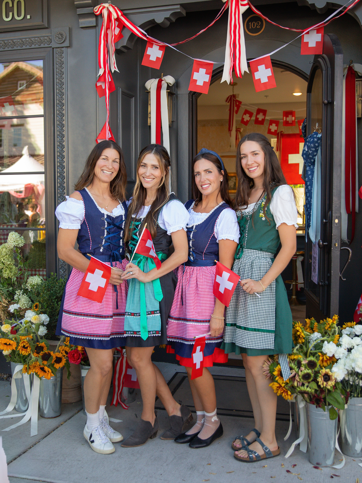 Flower Bar Co. staff wearing traditional Swiss dirndl dresses and holding small Swiss flags. The flower shop Midway storefront is decorated with Swiss flags, red-and-white ribbons, and flower arrangements on both sides.