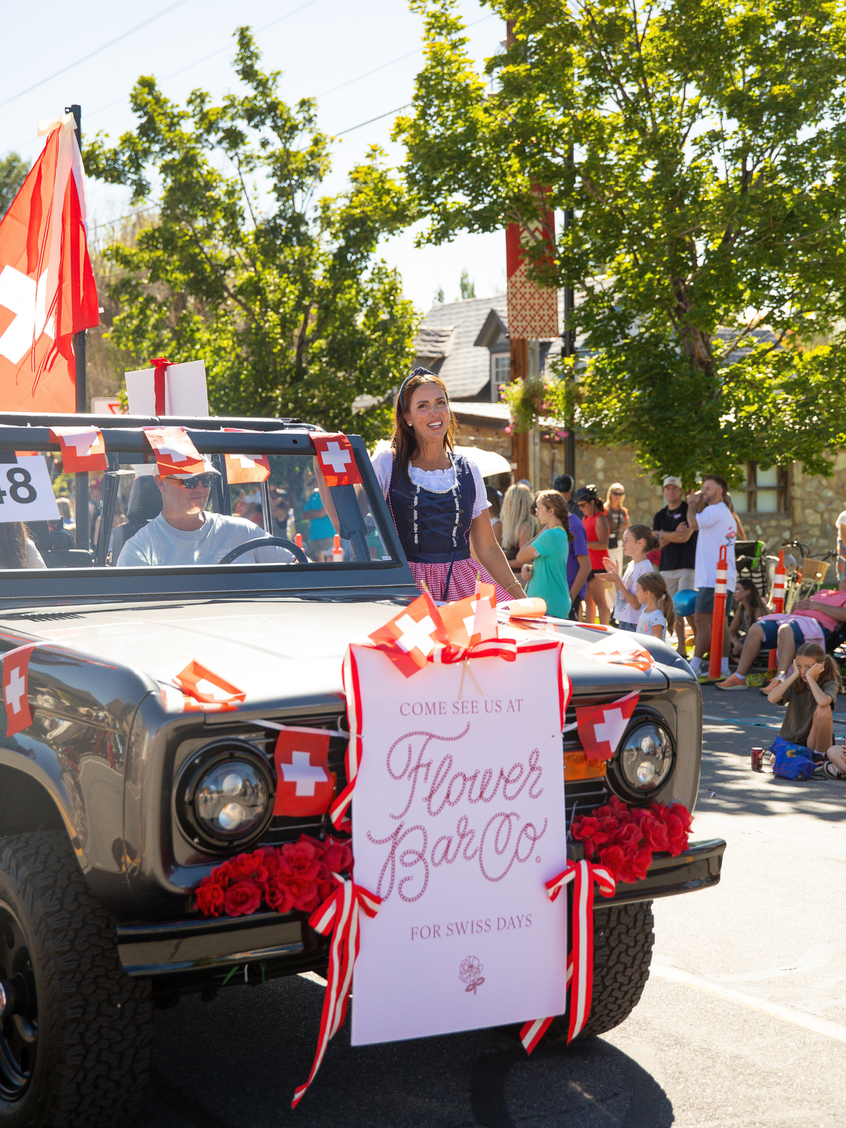 Flower Bar Co. foudner and florist Shawn Bailey in a traditional Swiss dirndl, stands beside a vintage Bronco decorated with Swiss flags and a large sign reading 'Come See Us at Flower Bar Co. For Swiss Days.