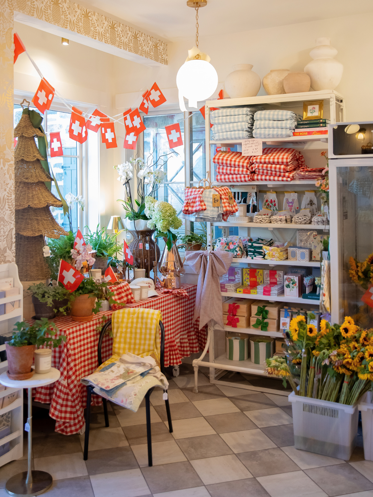 Flower Bar Co. Midway flower shop stocked with home decor, featuring red and white gingham checkered tablecloths, shelves of folded textiles, sunflowers, and Swiss flag bunting during Swiss Days.