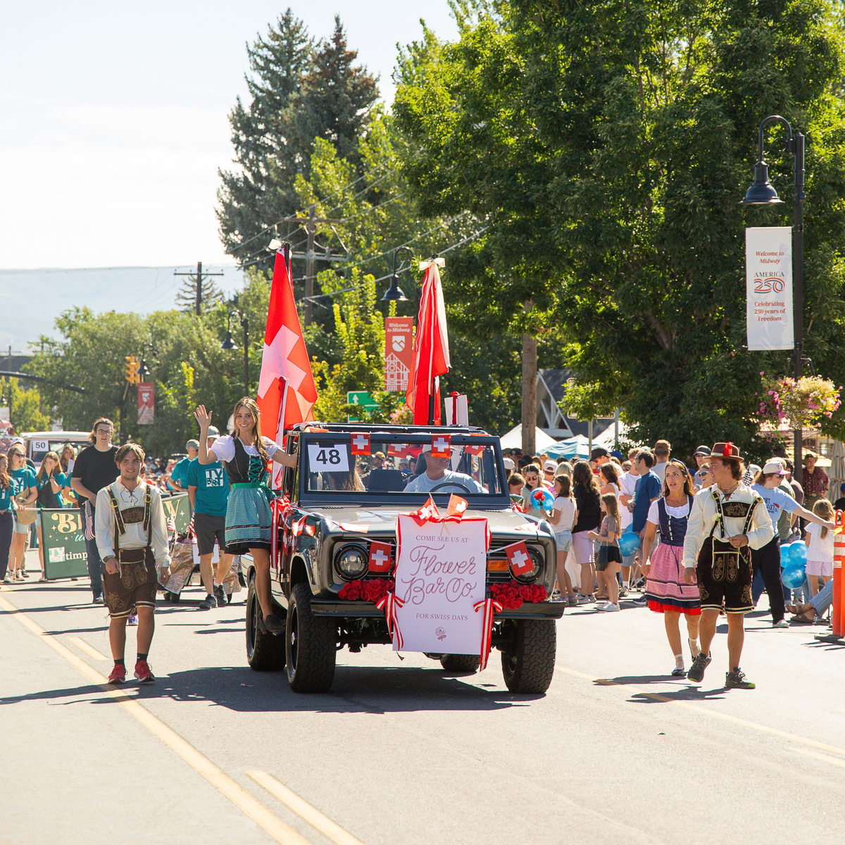 A vintage Brocno decorated with red bows and a 'Flower Bar Co.' sign drives down the Swiss Days parade, surrounded by spectators, trees, and Swiss flags.