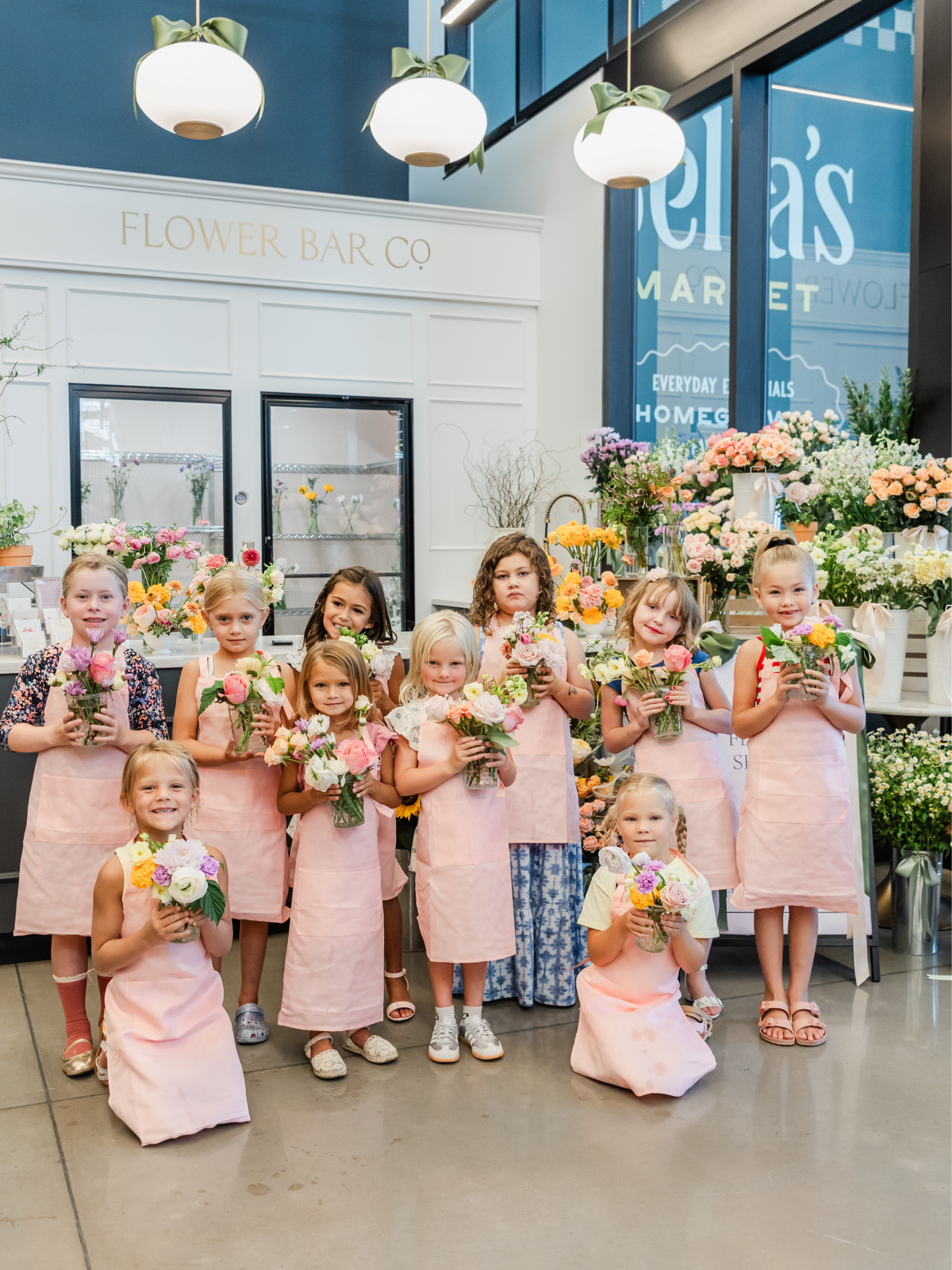 Young girls wearing matching pink aprons pose together in a flower shop, each holding a small bouquet of flowers. 