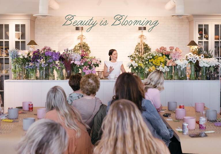Florist Shawn Bailey hosts a flower-arranging class among large displays of colorful flowers, speaking to an audience of women at a floral workshop