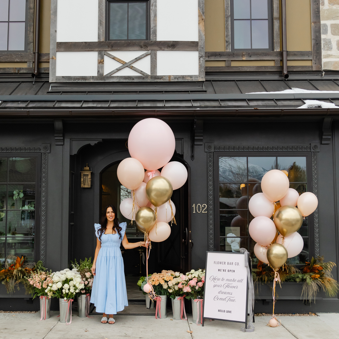 Florist Shawn Bailey stands in front of the Midway shop, holding a cluster of blush pink and metallic gold balloons. Two matching balloon arrangements sit on either side, with flower arrangements and a sign reading 'Flower Bar Co' at the bottom.