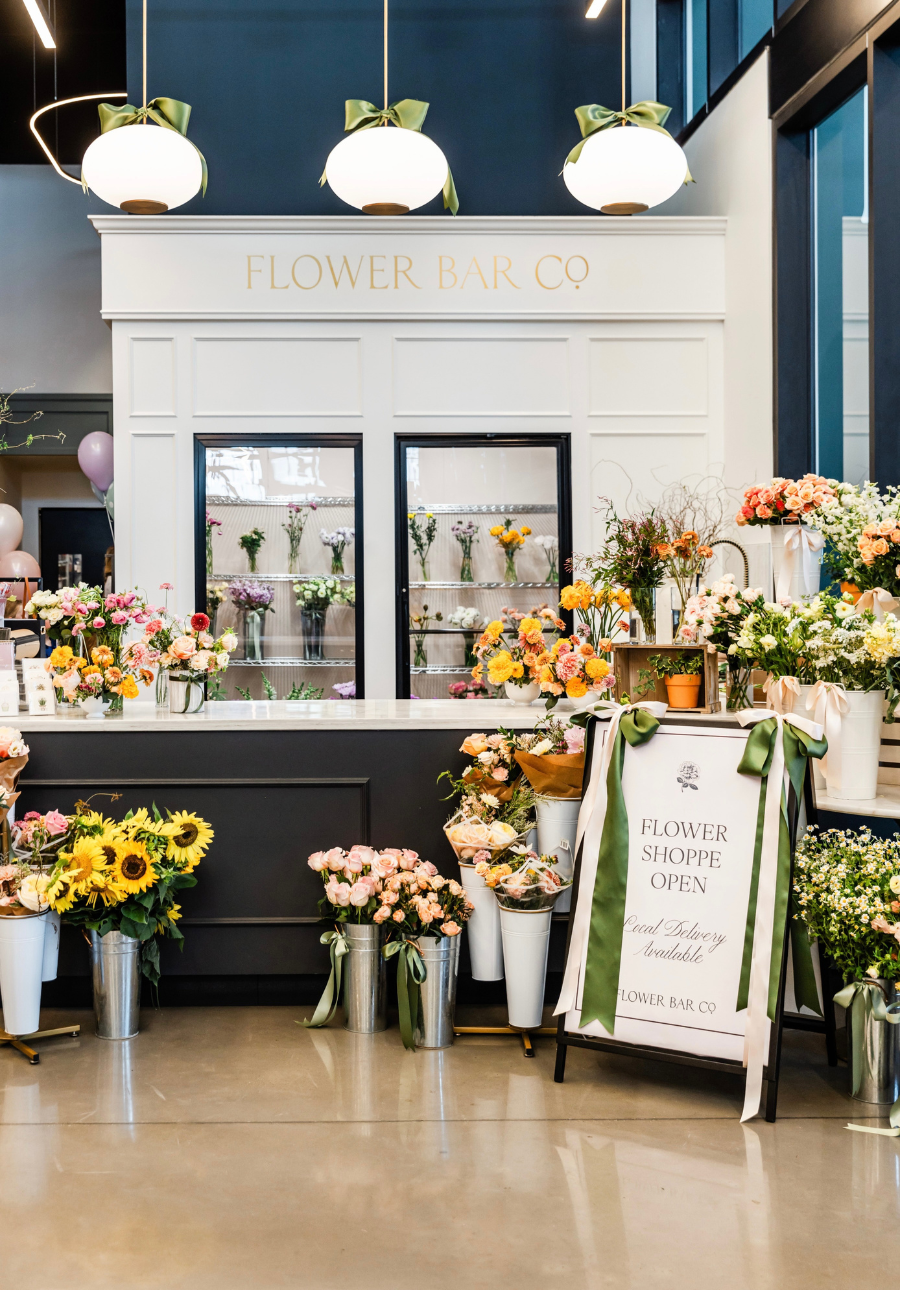 A bright flower shop interior with a white counter labeled 'FLOWER BAR CO,' multiple arrangements of sunflowers, roses, and other blooms
