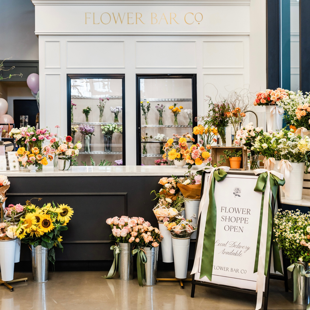 A Utah flower shop storefront with the words 'FLOWER BAR CO' above two display windows, surrounded by bouquets, flower arrangements, and a freestanding sign that reads 'FLOWER SHOPPE OPEN' with 'Flower Delivery Available' and 'FLOWER BAR CO' at the bottom.