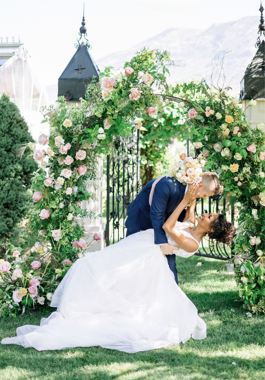 A bride in a white wedding gown and a groom in a navy suit under a lush green and pink floral arch in Utah