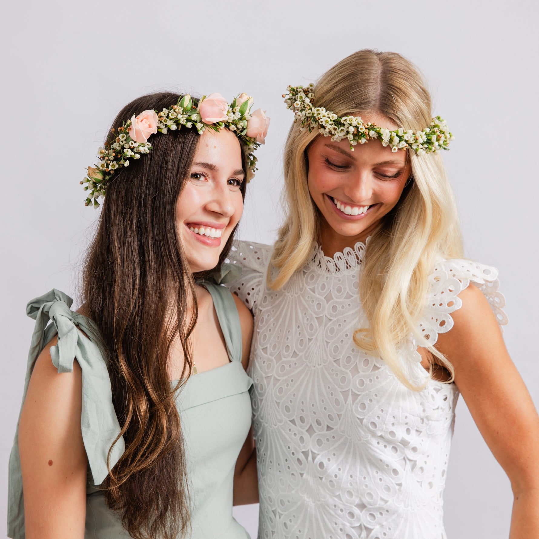 Two women smiling and wearing floral crown headbands, one in a sage green dress and the other in a white eyelet dress, against a light gray studio background.