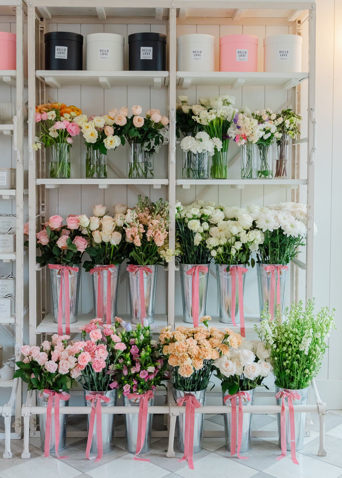 Flower Bar Co shelving unit filled with bouquets of pink, peach, white, and yellow flowers, with several metal buckets tied with pink ribbon.