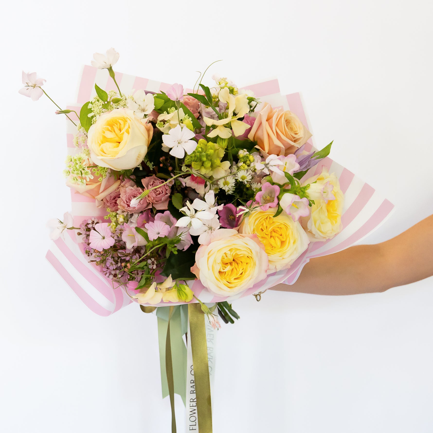 A vibrant floral bouquet featuring yellow and peach roses, pink carnations, white flowers, and green foliage, wrapped in pink striped paper with a green ribbon.