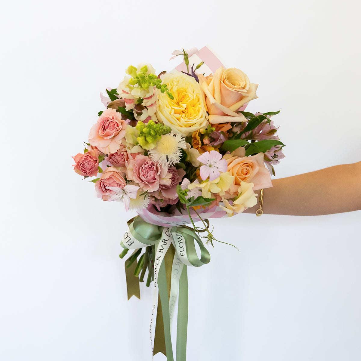 A pastel-colored flower bouquet featuring roses, chrysanthemums, and green foliage, wrapped in a light pink paper sleeve with a sage green ribbon. 