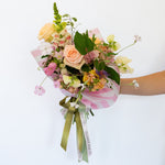 A fresh flower bouquet wrapped in pink and white striped paper with a green ribbon printed with 'Flower Bar Co'. The bouquet features a peach rose, yellow carnations, pink blossoms, and green foliage .
