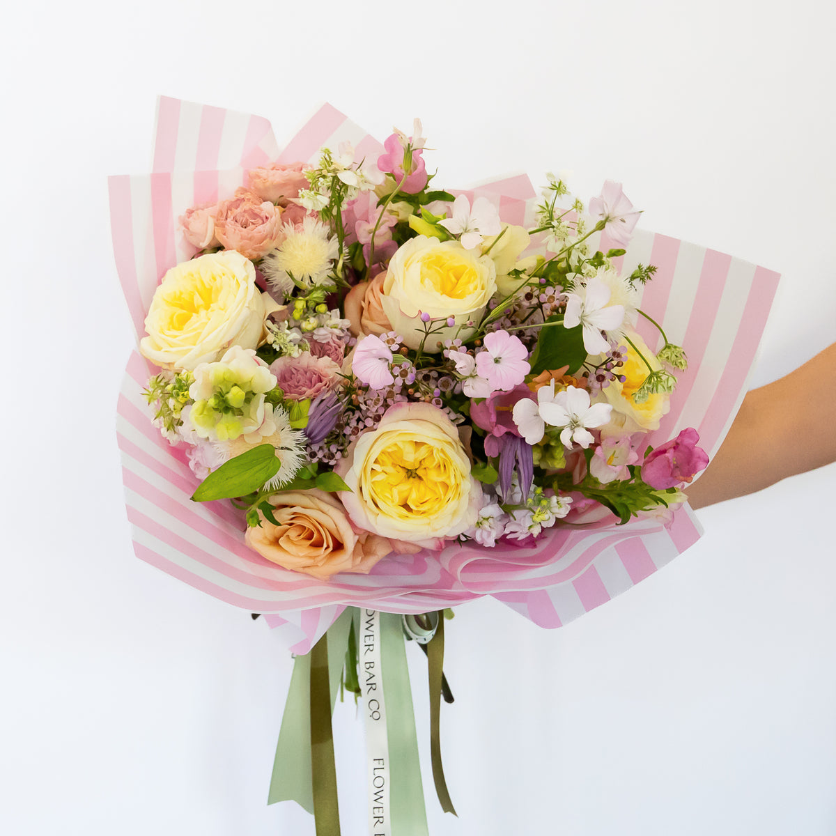 A vibrant hand-tied bouquet of fresh flowers: yellow and peach roses, pink carnations, purple stock, and green foliage, wrapped in pink and white striped paper, tied with a ribbon from 'Flower Bar Co'.