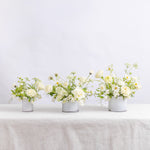 Three small white vases filled with a white floral arrangement featuring roses, daisies, and green foliage.