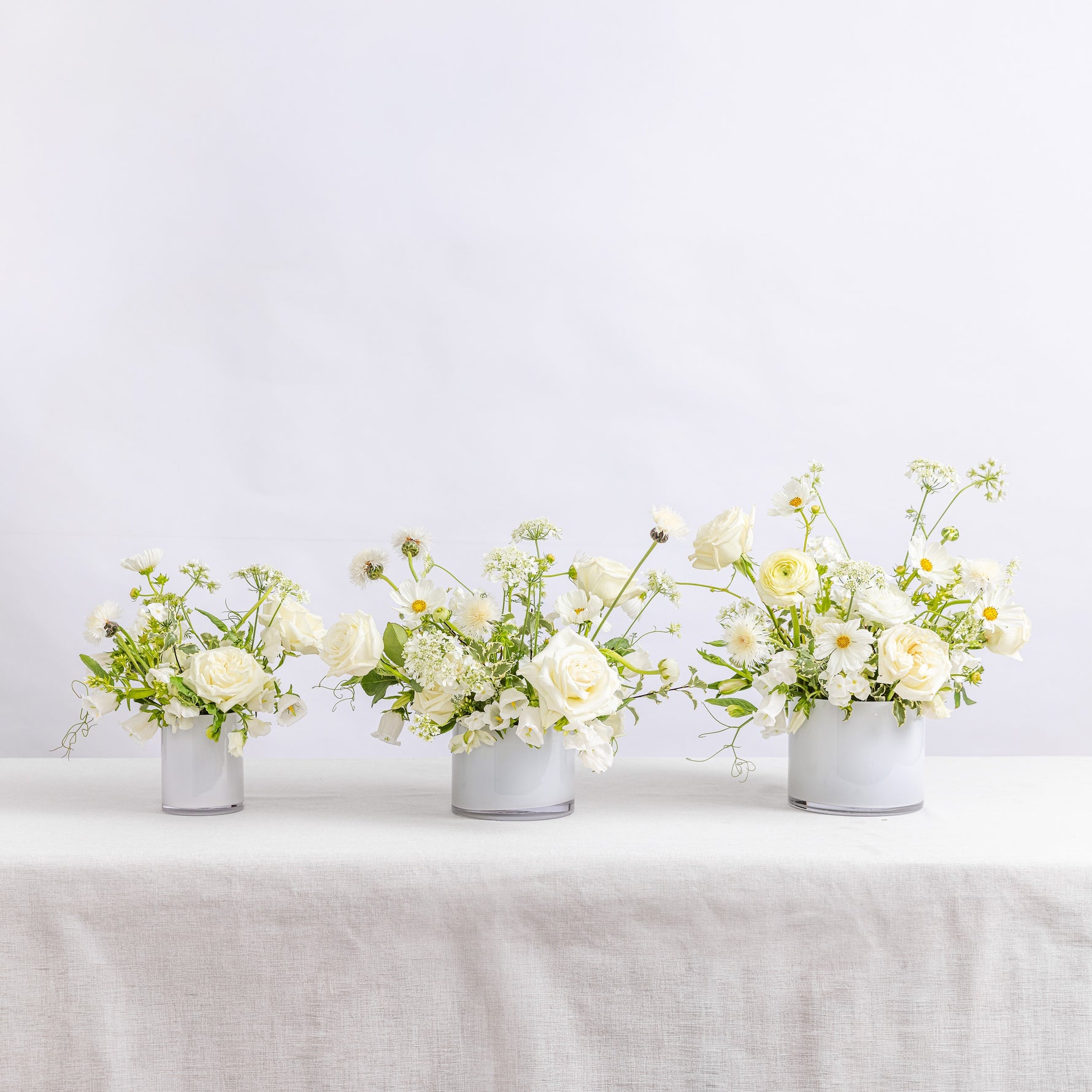 Three small white vases filled with a white floral arrangement featuring roses, daisies, and green foliage.