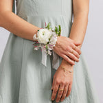A woman wearing a white floral wrist corsage with green leaves and white ribbons, for weddings or school dances.