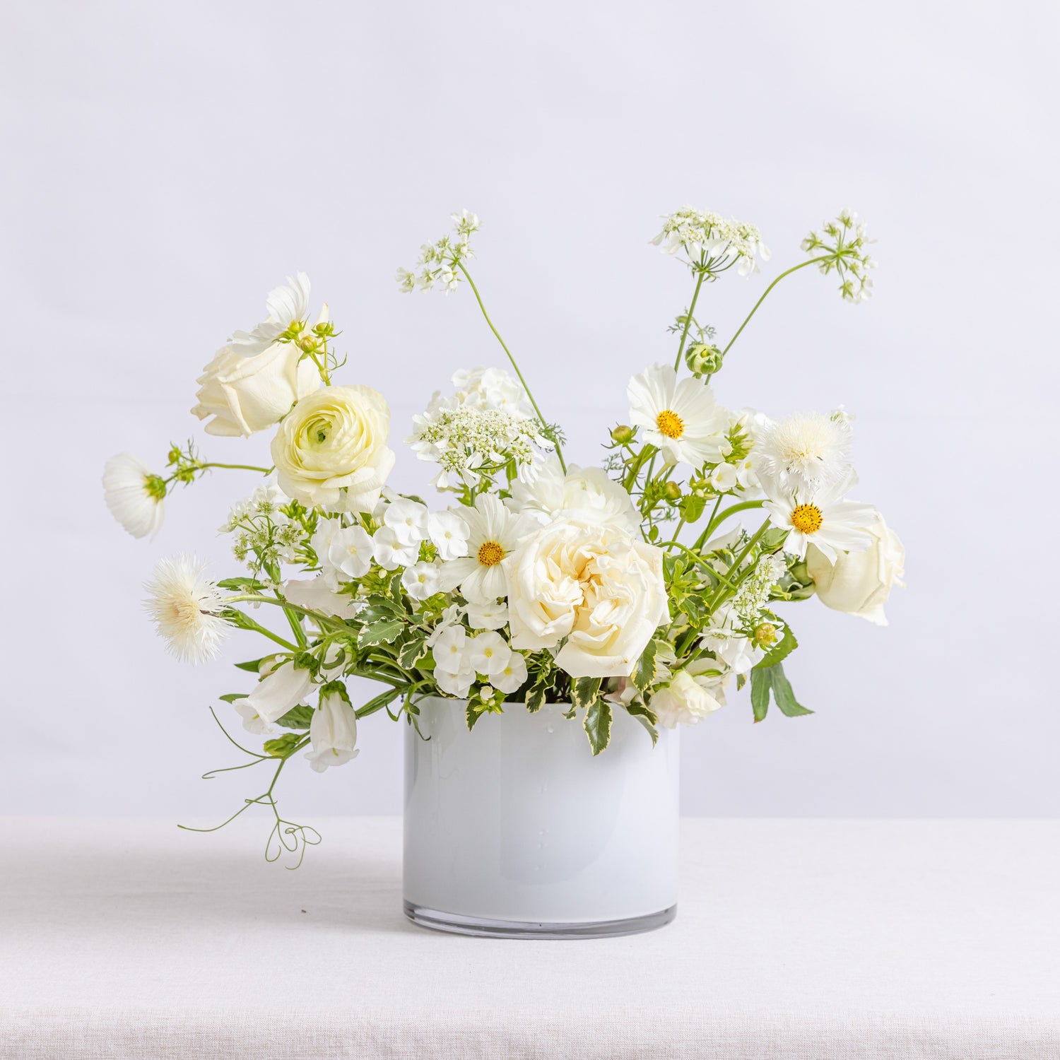 A white vase filled with a bouquet of white roses, daisies, and green foliage.