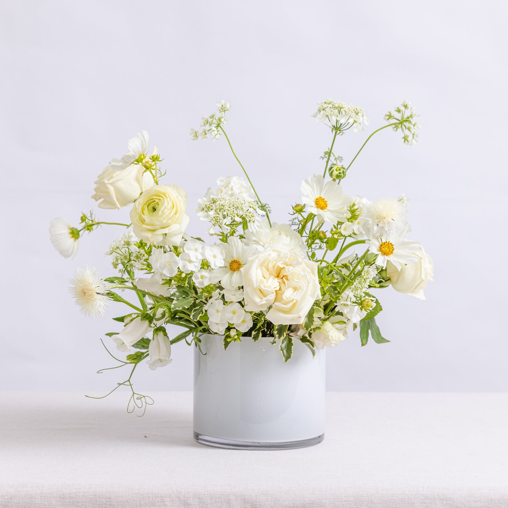 A white vase filled with a bouquet of white roses, daisies, and green foliage.