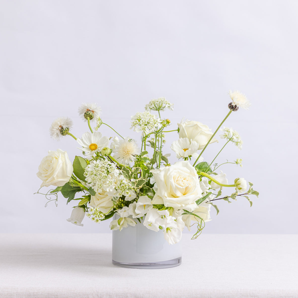 A floral arrangement of white roses, daisies, and small white flowers with green stems, presented in a white vase.