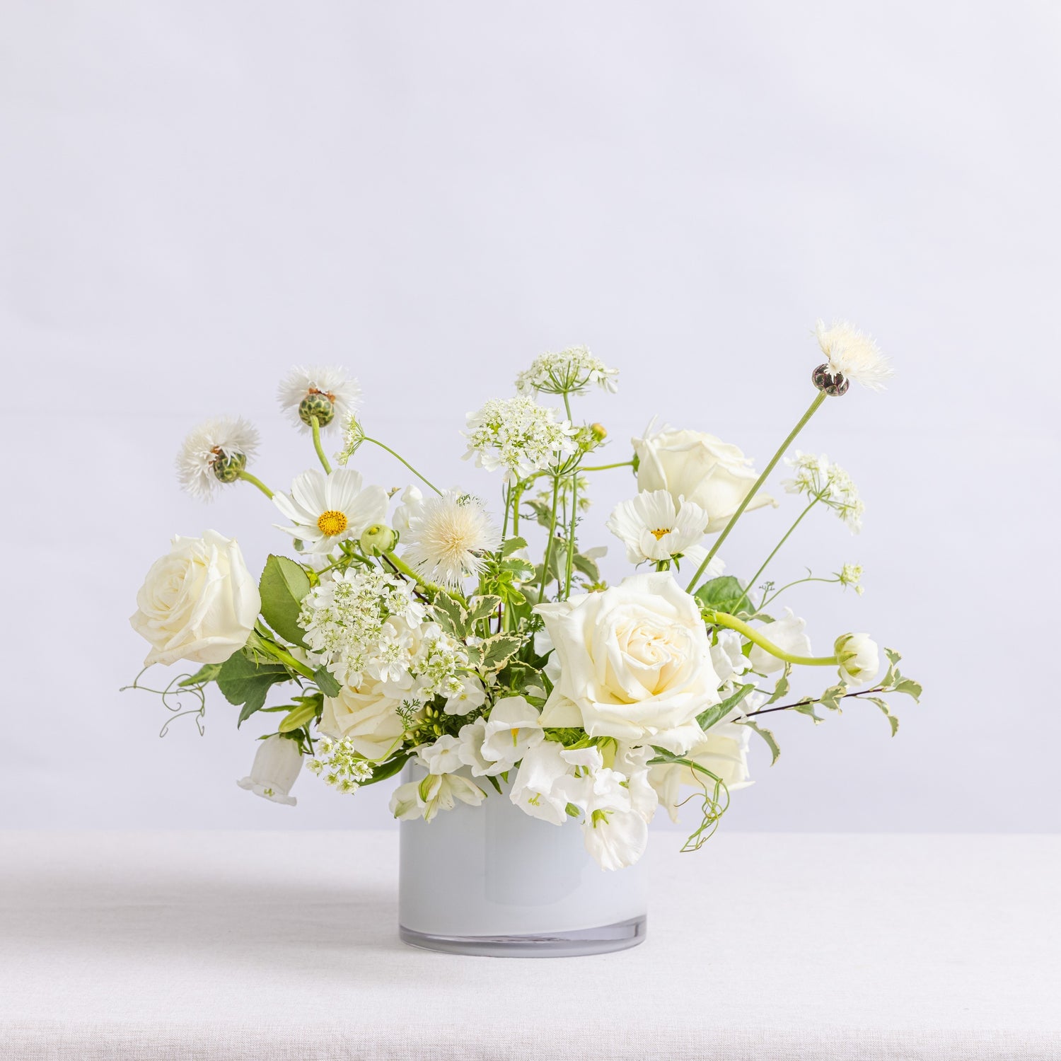 A floral arrangement of white roses, daisies, and small white flowers with green stems, presented in a white vase.