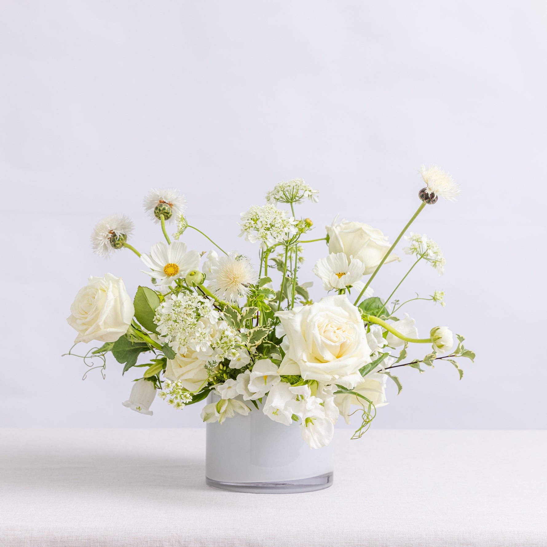 A floral arrangement of white roses, daisies, and small white flowers with green stems, presented in a white vase.