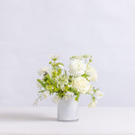 A small floral arrangement of white roses, daisies, and green foliage in a simple white vase