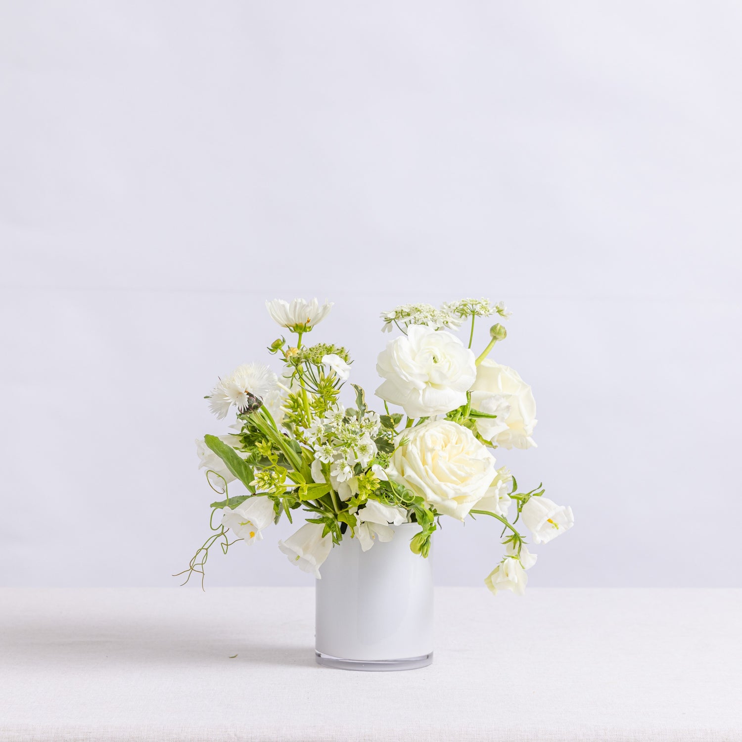 A small floral arrangement of white roses, daisies, and green foliage in a simple white vase