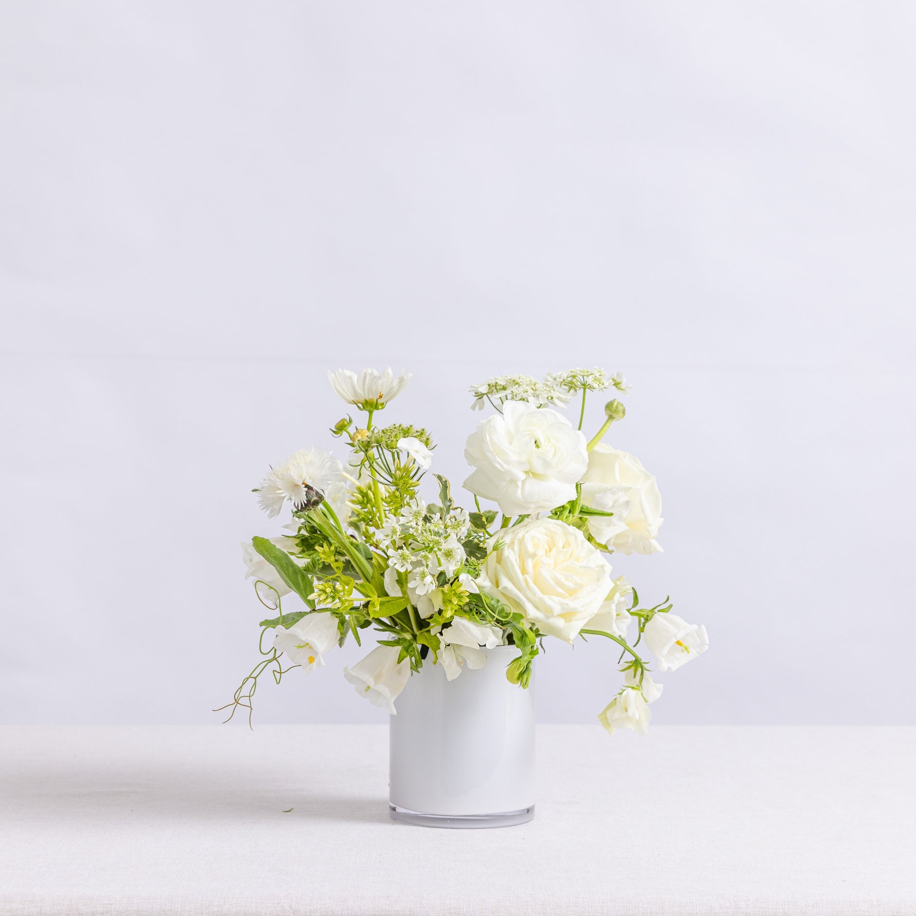 A small floral arrangement of white roses, daisies, and green foliage in a simple white vase
