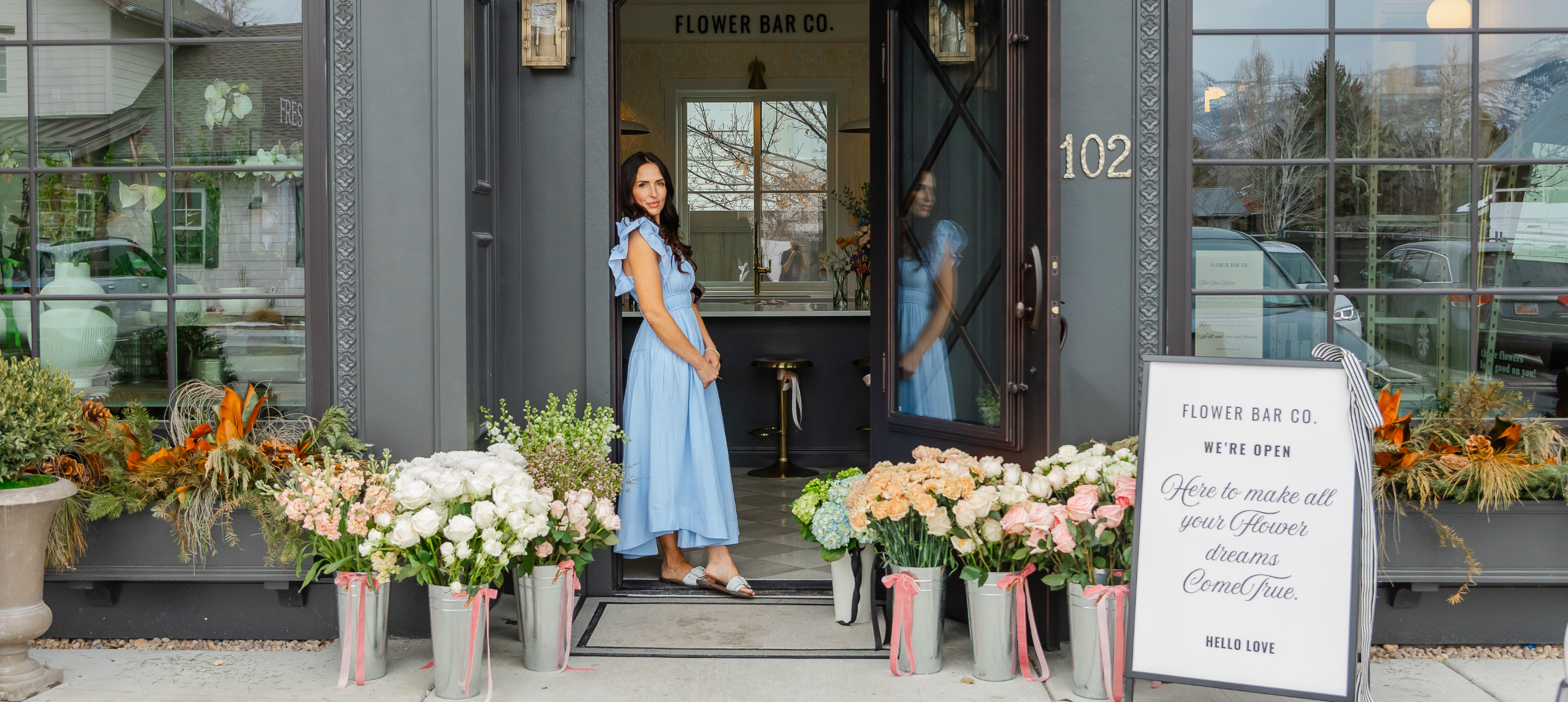 Owener Florist Shawn Bailey stands in the doorway of a flower bar shop, with potted flowers arranged along the front steps and a sign reading 'Flower Bar Co.'