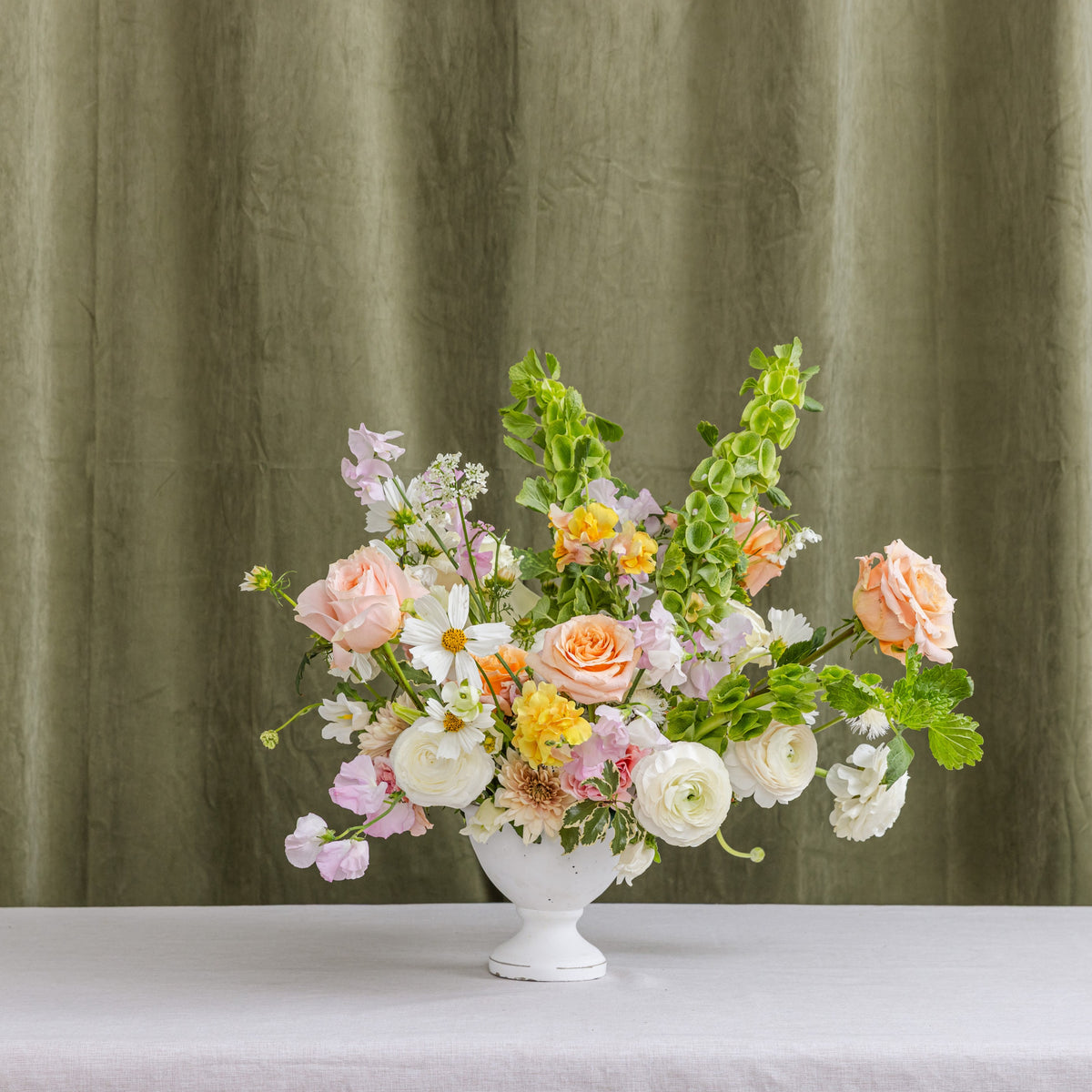 A floral arrangement in a white pedestal vase, featuring peach roses, white ranunculus, daisies, and green foliage.