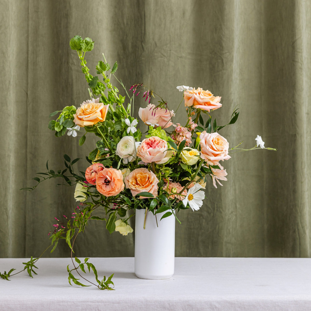 A tall floral arrangement of peach, pink, cream, and white flowers with green foliage in a white cylindrical vase.