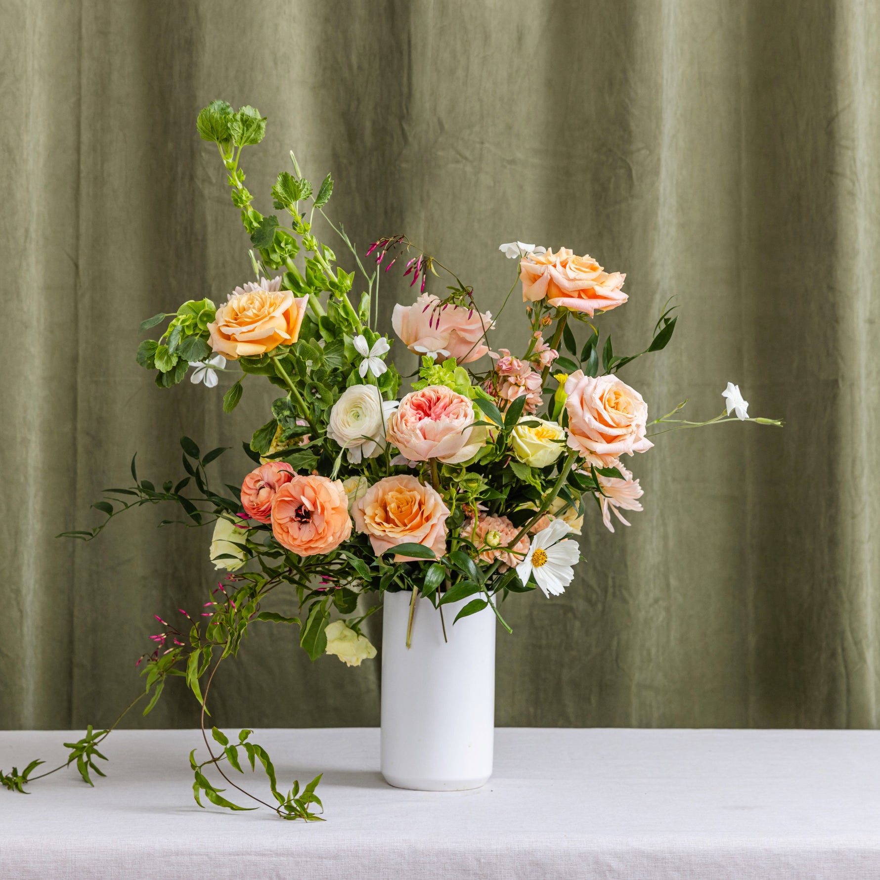 A tall floral arrangement of peach, pink, cream, and white flowers with green foliage in a white cylindrical vase.