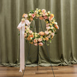A circular floral wreath in soft peach, pink, cream, and yellow tones with green foliage.